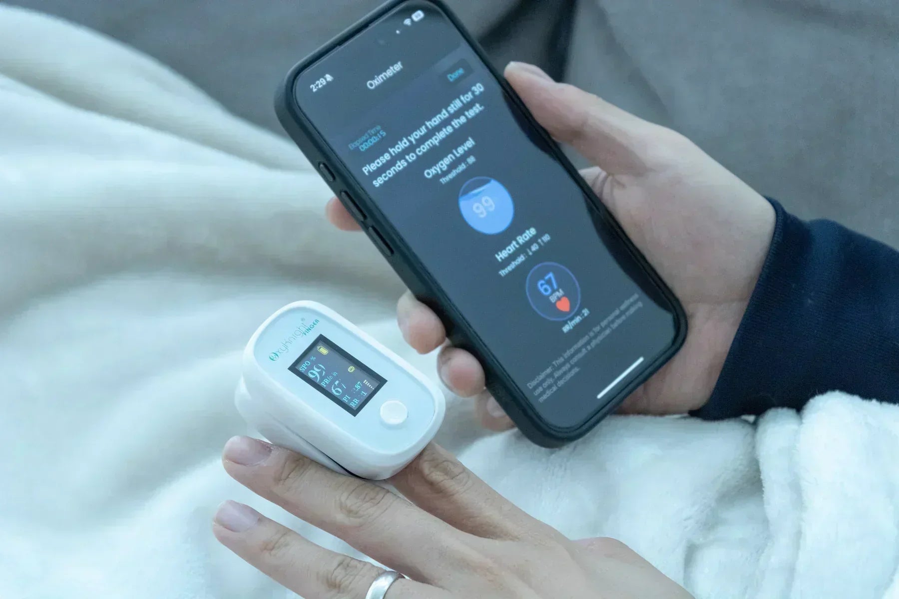 A person uses a fingertip pulse oximeter connected to a smartphone app to monitor oxygen saturation and pulse rate in real time.