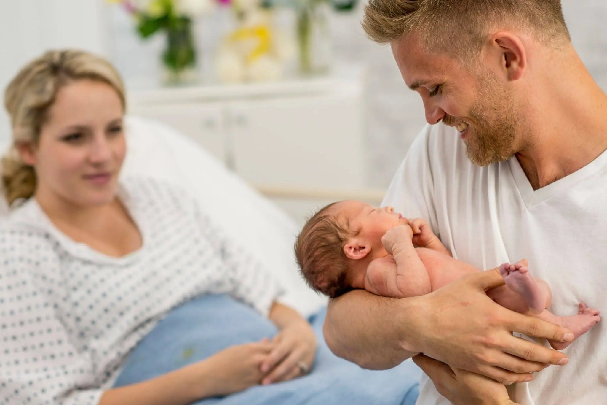 Father holding his newborn baby while the mother watches in a hospital room following routine newborn screenings.