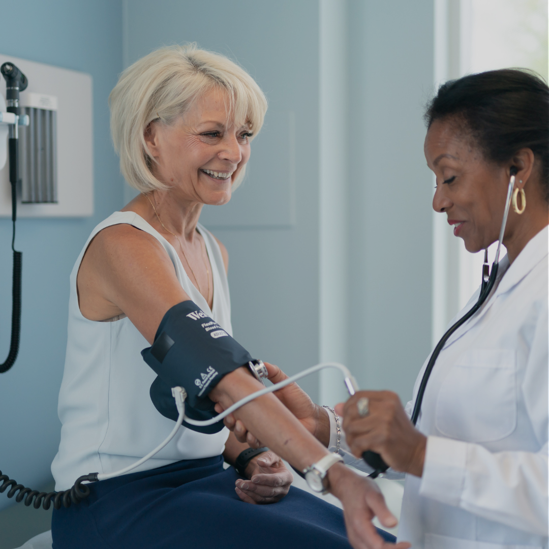 A woman is smiling in her doctors office while her doctor takes her blood pressure with a health monitoring device